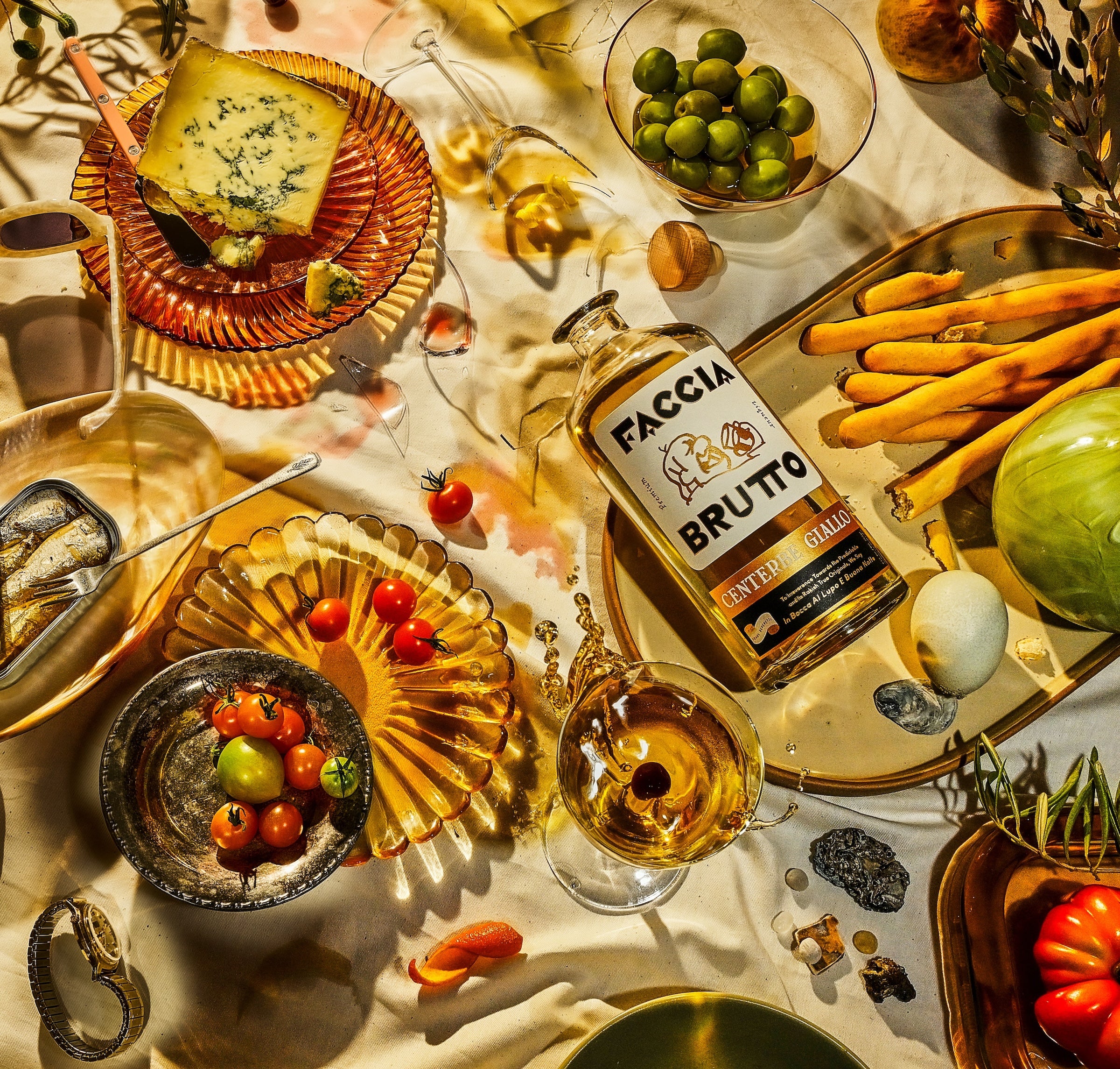 Tablescape with Faccia Brutto Centerbe Giallo bottle, cheese, olives, tomatoes, glasses, breadsticks