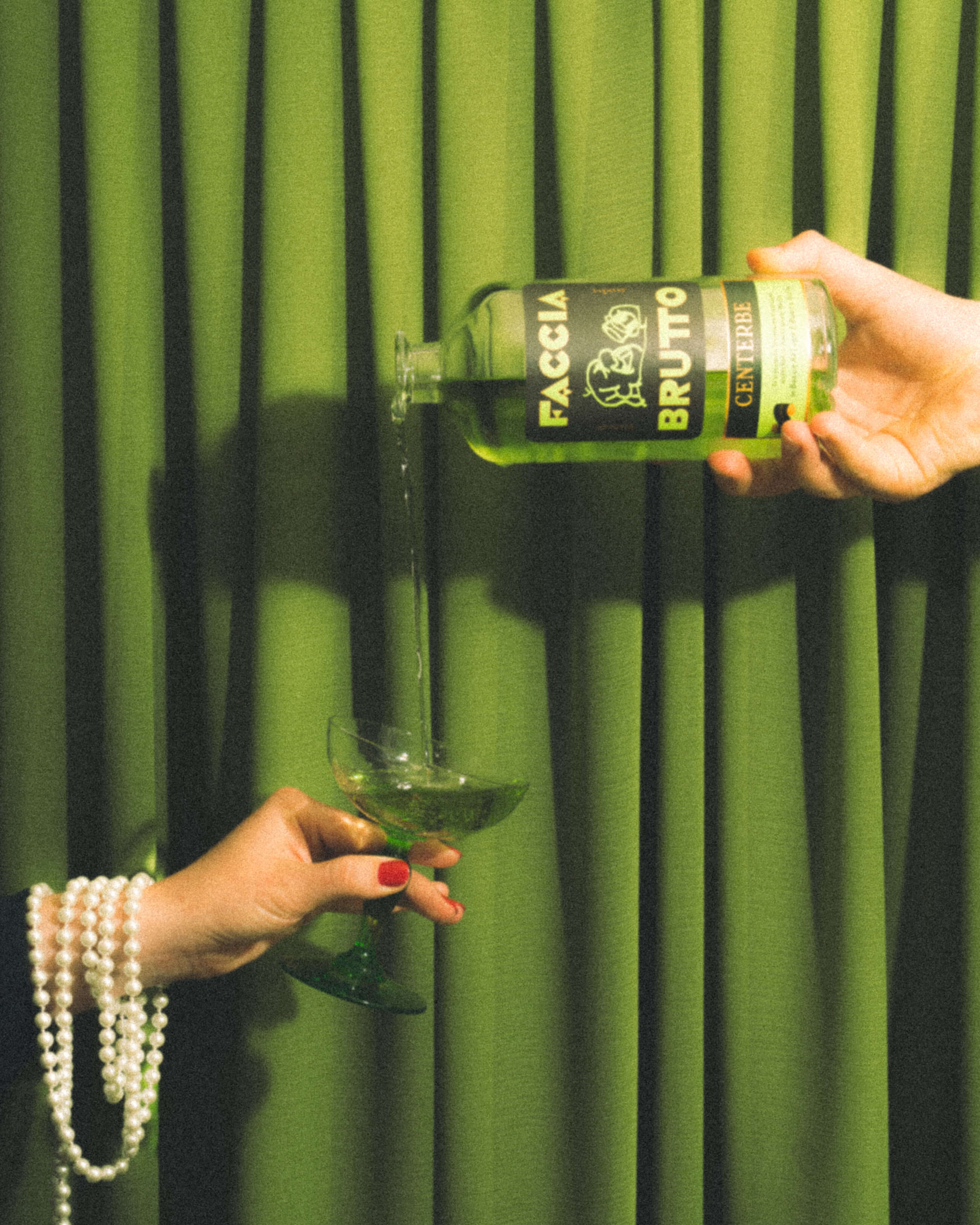 Hand pouring Faccia Brutto Centerbe bottle into a couple glass in front of green curtain