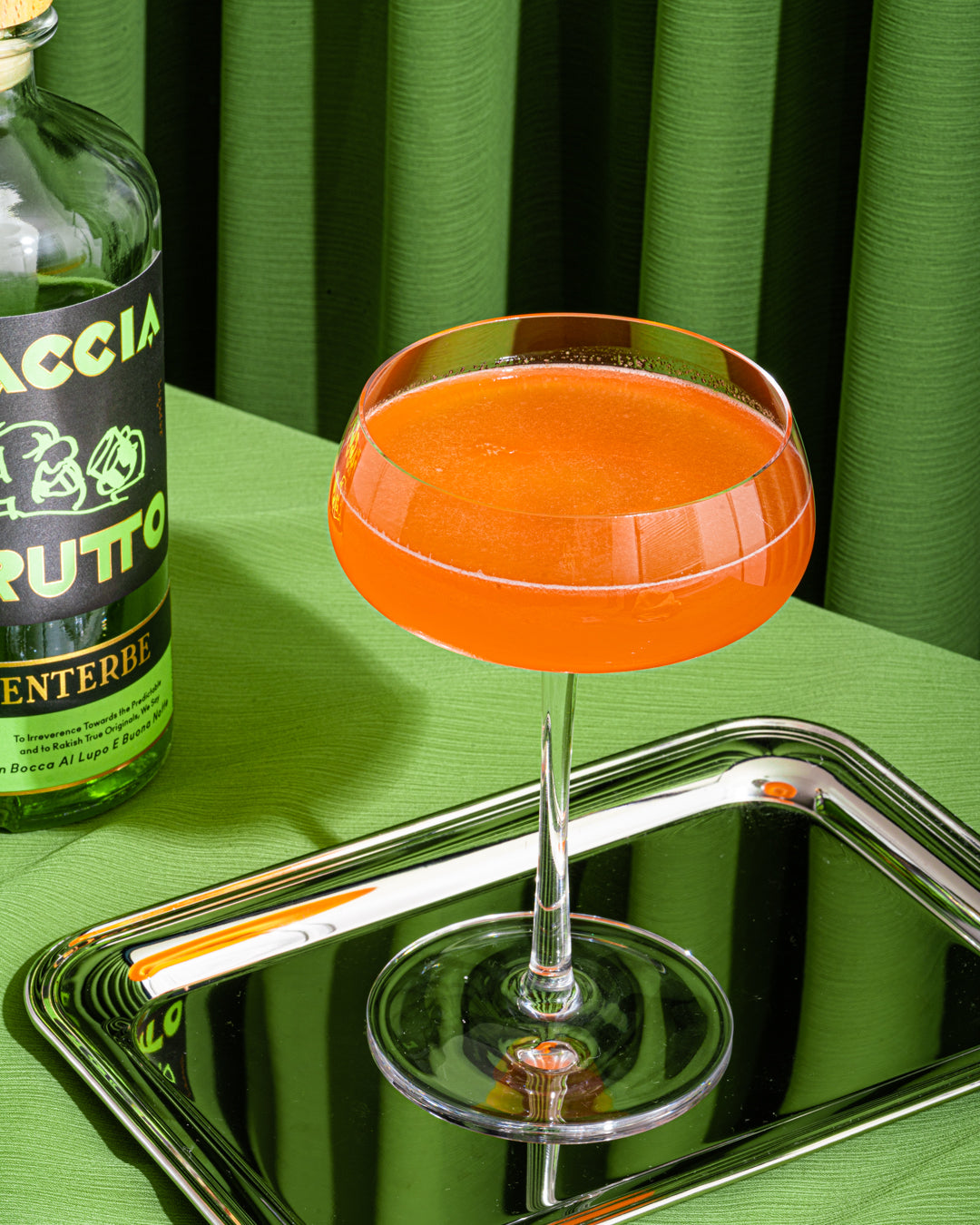 Bright orange cocktail on shiny silver tray with bottle of Faccia Brutto Centerbe in front of green curtain