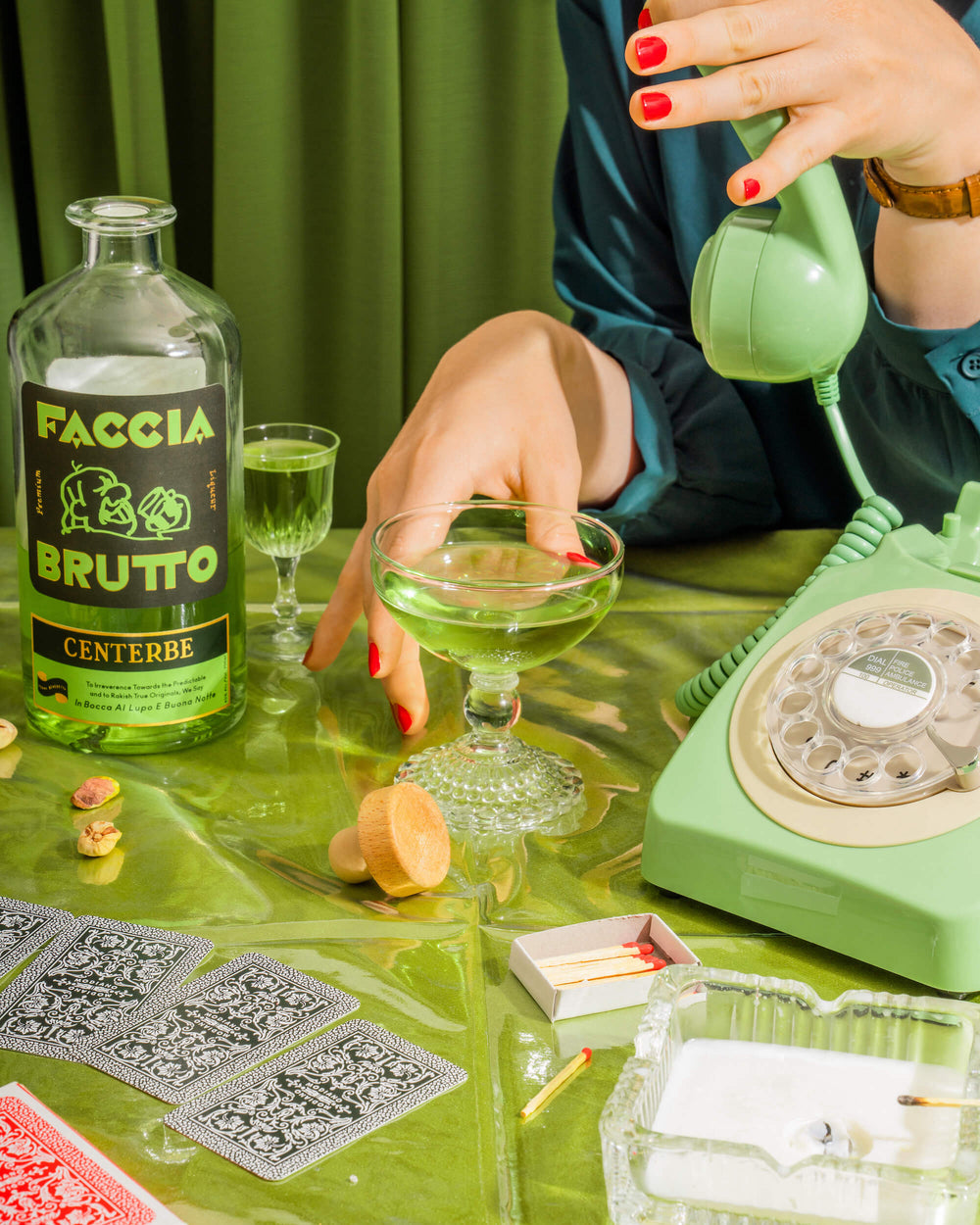 Tablescape with playing cards, matches, ashtray, green rotary phone, hands with red painted nails, cocktail glasses and a bottle of Faccia Brutto Centerbe