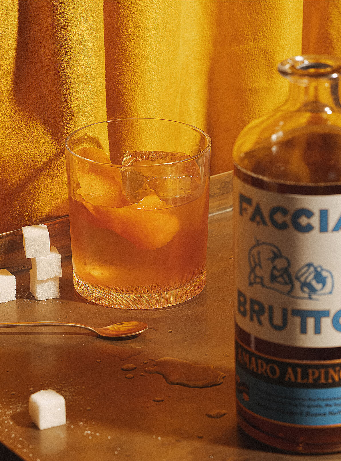 Cocktail with sugar cubes, spoon and out of focus Faccia Brutto bottles in front of yellow curtain