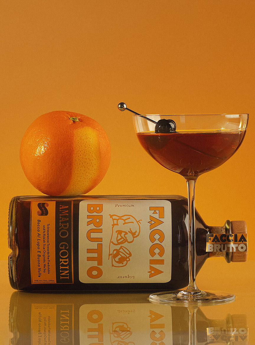Bottle of Faccia Brutto on its side, orange with missing section of peel, and cocktail in tall coupe glass with dark cherry garnish