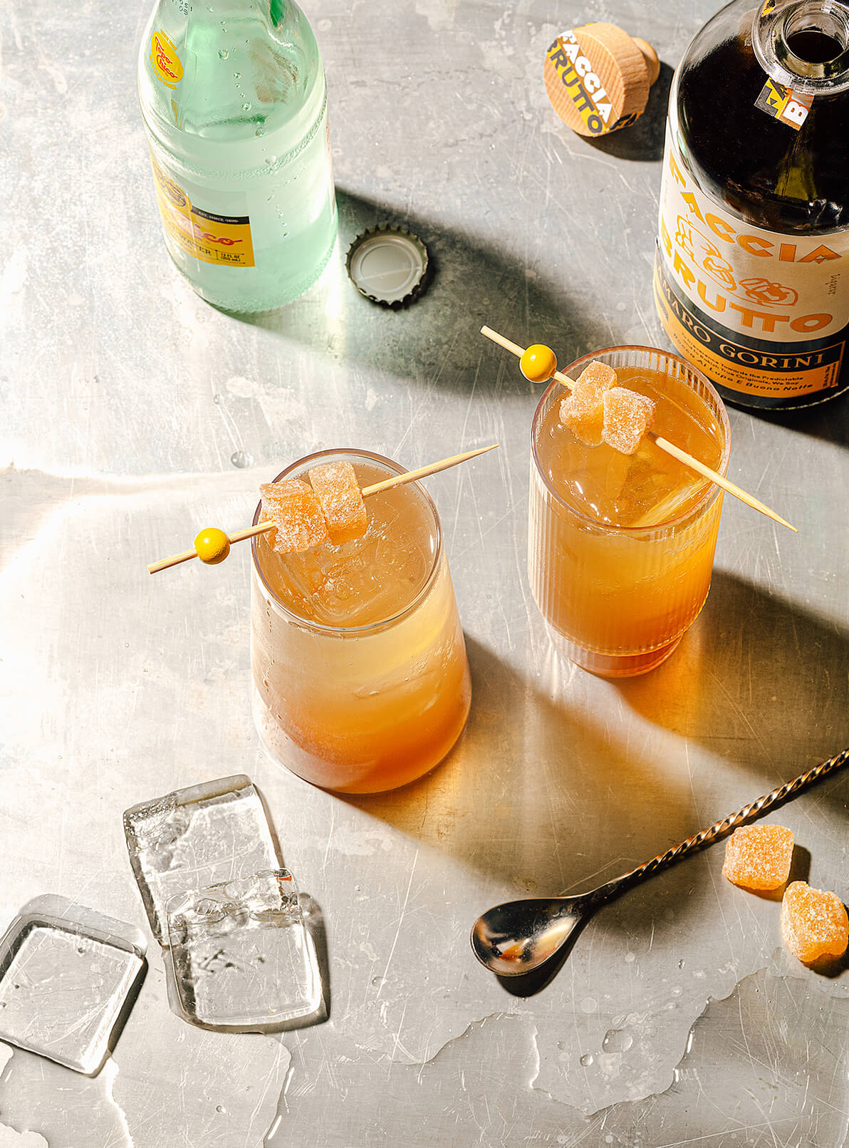 Two tall cocktails with ice and garnishes, bottle of Topo Chico and bottle of Faccia Brutto Amaro Gorini