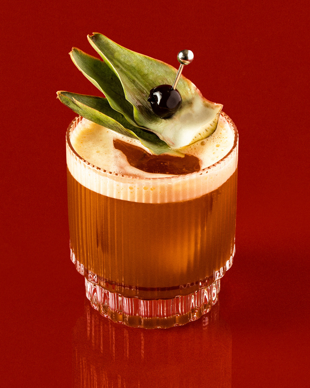 Cocktail glass with pineapple leaves, dark cherry and foam on glossy red background